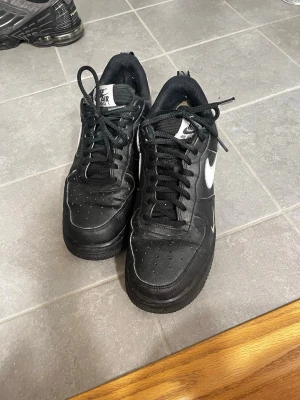 Nike Air Force 1 svart/vit sneaker - Säljer ett par klassiska Nike Air Force 1 sneakers i svart skinn med vita detaljer och swoosh på sidan. Skorna har snörning, rund tå och platt sula. Perfekta för dig som gillar streetwear och vill ha en tidlös look.