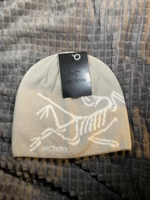 Beige mössa från Arc'teryx - Säljer en beige mössa från Arc'teryx med deras ikoniska fossil-logga i vitt. Mössan är ribbstickad och har en clean look som passar till det mesta. Tillverkad i 100% akryl vilket gör den mjuk och skön att bära.