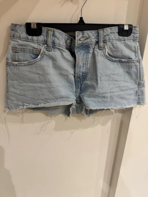 Ljusblå jeansshorts från 157 - Ljusblå jeansshorts från 157 med råa, fransiga kanter och klassisk femficksdesign. Shortsen har bälteshällor och knappgylf. Perfekta för varma dagar och ger en avslappnad vibe.