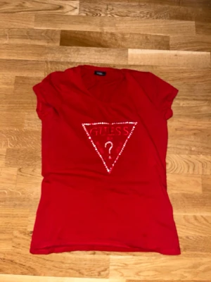 Röd t-shirt från Guess med paljetter - Snygg röd t-shirt från Guess med det ikoniska trekantsloggan framtill, dekorerad med glittriga paljetter. T-shirten har korta ärmar och en klassisk passform. Perfekt för dig som vill sticka ut lite extra.