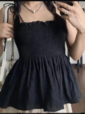 Svart smockad linnetopp med volang - Säljer en svart linnetopp med smockad överdel och volangig nederdel. Toppen har tunna axelband som knyts på axlarna och en söt rynkad kant upptill. 