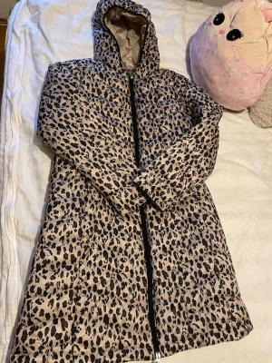 Leopardmönstrad pufferjacka med huva - Säljer en lång, quiltad pufferjacka från mywear woman i storlek S. Jackan har ett snyggt leopardmönster i beige och svart, dragkedja framtill och en stor huva. Perfekt för dig som vill sticka ut i vinter och hålla dig varm.