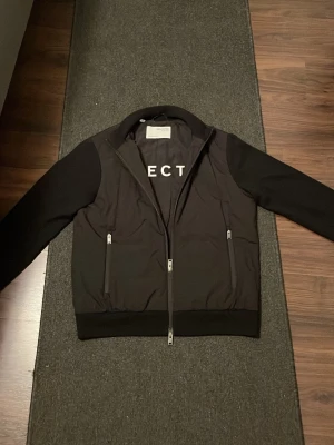 Svart Cardigan från Selected Homme - Säljer en svart cardigan från Selected Homme med minimalistisk design. Jackan har två sidofickor med dragkedja, ribbade muddar och krage samt diskret logga på insidan. Materialet är en mix av polyester och bomull för en skön känsla. 