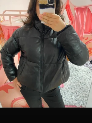 Svart pufferjacka i skinnimitation - Säljer en svart pufferjacka med hög krage och dragkedja framtill. Jackan har en oversized passform och är gjord i skinnimitation som ger en snygg och modern look. Perfekt för kalla dagar och enkel att styla till olika outfits.