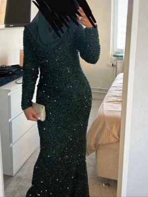 Mörkgrön glittrig långklänning - Säljer en mörkgrön långklänning med massor av glittrande paljetter. Klänningen har lång ärm och en figurnära passform som framhäver siluetten. Perfekt för bal eller festliga tillfällen där du vill sticka ut och glänsa. Endast andvänd en gång!!