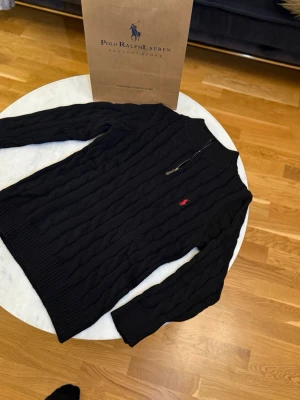 Svart stickad tröja Polo Ralph Lauren - Säljer en svart kabelstickad tröja från Polo Ralph Lauren med halv dragkedja och röd broderad logga på bröstet. Tröjan har ribbade muddar, hög krage och är långärmad. Perfekt för dig som vill ha en klassisk och stilren look.
