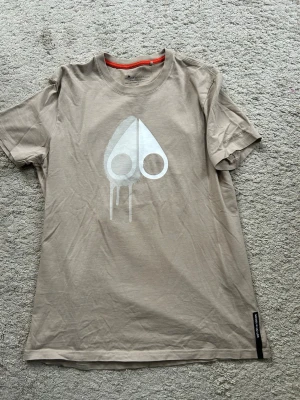 Beige Moose Knuckles t-shirt med tryck - Beige t-shirt från Moose Knuckles som är köpt på Zgander i Helsingborg i storlek Medium. Tröjan är använd fåtalet gånger och är i mycket bra skick.