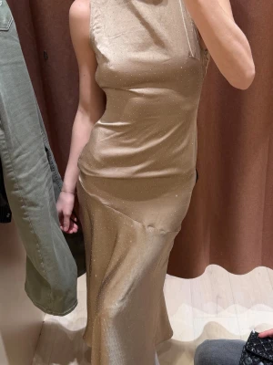 Beige långklänning med öppen rygg - Supersnygg beige långklänning i glansigt material med små diskreta prickar. Klänningen har en elegant öppen rygg med draperad detalj och är ärmlös. Sitter figurnära upptill och faller snyggt längs kroppen. Perfekt för dig som vill sticka ut med en stilren look.