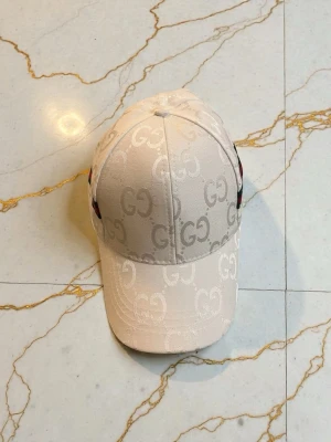 Vit Gcci keps med GG-mönster - Säljer en beige keps från Gcci med ikoniskt GG-mönster över hela kepsen. Klassisk böjd skärm och snygg detalj med röd-grön rand på sidan. Tillverkad i ett glansigt material som ger en lyxig känsla. Perfekt accessoar för att lyfta din outfit.