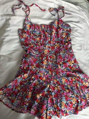 Blommig kortklänning med knytband - Kortklänning med färgglatt blommönster. Klänningen har tunna axelband med knyt, markerad midja och med insydd skjorts. Storlek (M). Skriv via dm vid funderingar 💬🌺