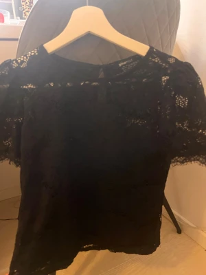 Svart spetsblus från Gina Tricot - Säljer en svart blus i spets från Gina Tricot. Blusen har korta ärmar, rund halsringning och knäppning bak i nacken. Spetsen har ett blommigt mönster och ger en snygg transparent effekt. Perfekt att styla med jeans eller kjol.