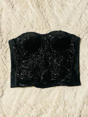 Svart glittrig paljettkorsett - Säljer en svart korsett topp med massor av svarta paljetter på framsidan som ger ett riktigt glittrigt intryck. Toppen är axelbandslös och har en stadig form med vadderade kupor och hakar i ryggen. Sidorna och baksidan är i blankt satinliknande material.