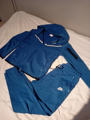 Blå Nike mjukisbyxor med logga - Säljer ett par blå mjukisbyxor från Nike med vit logga på benet. Byxorna har dragsko i midjan och muddar vid bensluten. Tillverkade i mjukt bomullsmaterial, perfekta för chill eller träning. Matchande hoodie finns också om du vill köra hela setet. Tröjan 147 till 158 och byxorna är 158 till 170