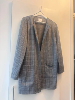 Rutig kavaj från Vila, storlek L - Snygg rutig kavaj från Vila i svart, grått och vitt. Rak passform med långa ärmar och två fickor framtill. Perfekt för dig som vill ha en stilren och trendig look. Klassisk modell utan knappar och med mjukt tyg.