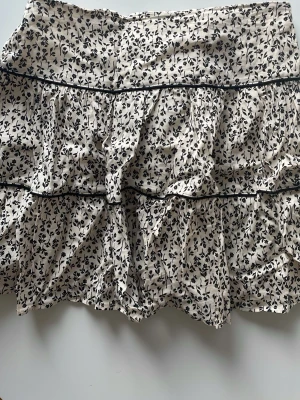 Vit blommig kortkjol med volanger - Supersöt kortkjol i vitt med svart blommönster och volangdetaljer. 