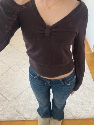 Brun figursydd stickad tröja - Mörkbrun finstickad tröja med v-ringning och snygg detalj framtill. Långa ärmar med knappar vid ärmsluten och tajt i midjan. Hör av er vid funderingar!💕