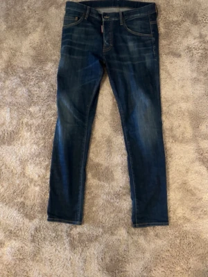 Mörkblå  jeans från Dsquared2 - Säljer ett par mörkblåa jeans från Dsquared2 med snygga slitningar och klassiska fem fickor. Jeansen har en röd DSQ2 Brothers-lapp bak och detaljerade sömmar. Materialet är bomull med lite elastan för extra stretch och bekvämlighet.