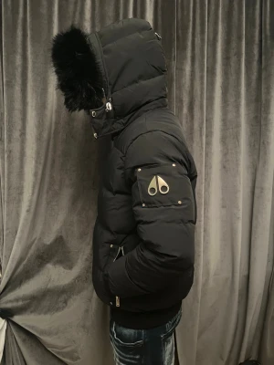  Moose Knuckles - Säljer en svart pufferjacka från Moose Knuckles med gulddetaljer och stor huva med svart fuskpäls. Jackan har flera dragkedjor, ribbade muddar och ikonisk logga på ärmen. Insidan har snygga band med logotyp och innerfickor. Perfekt för kalla dagar.
