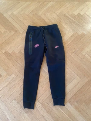 Syna world joggers - KOMMER EJ MED LAPPARNA⚠️.    Svarta mjukisbyxor från Nike i slim fit med taperad benform och regular längd. Byxorna har rosa Nike-logga och snörning i midjan samt en dragkedjeficka på sidan. Materialet är mjukt och bekvämt, perfekt för chill eller träning.