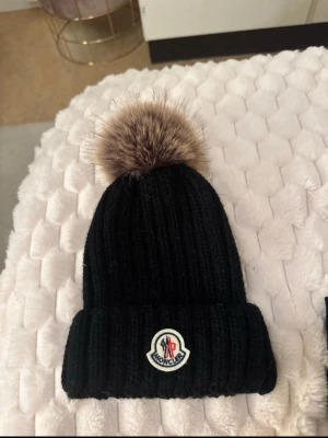 Svart Moncler mössa med pälsboll - Säljer en svart ribbstickad mössa från Moncler med stor fluffig pälsboll på toppen. 
