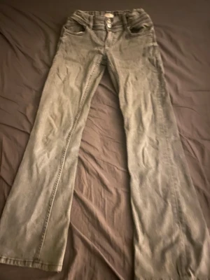 Grå bootcut jeans storlek 158 - Säljer ett par gråa bootcut jeans med hög midja och klassisk femficksdesign. Jeansen har breda ben och dubbla knappar i midjan för extra detalj. Perfekta för dig som gillar en retroinspirerad look.