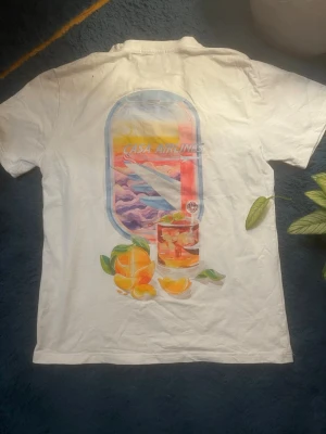 Casa Airlines t-shirt från Casablanca - Vit t-shirt från Casablanca med färgglad grafik på både bröstet och ryggen. Trycket visar ett flygplansfönster med solnedgång, drink och apelsiner samt texten 'Casa Airlines'. Skön passform och klassisk rund hals.