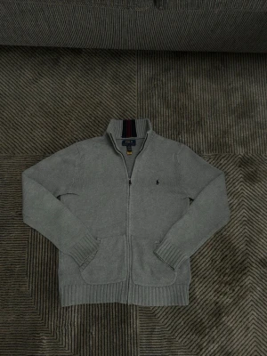 Grå stickad kofta från Polo Ralph Lauren - Säljer en grå stickad kofta från Polo Ralph Lauren med dragkedja framtill och två fickor. Hög krage med marinblå och vinröda ränder på insidan. Klassisk logga broderad på bröstet och ribbade muddar vid ärmslut och nederkant.