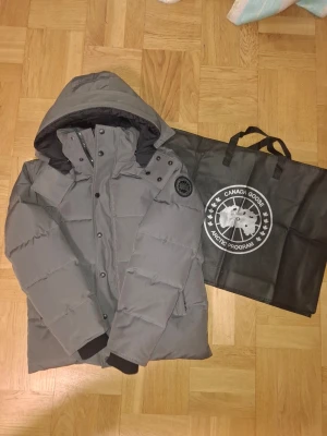 Grå Canada Goose - Säljer en grå pufferjacka från Canada Goose med huva och svarta detaljer. Jackan har quiltad design, dragkedja och knappar framtill samt logotyp på ärmen. Perfekt för kalla dagar och riktigt snygg till streetwear- stilen. Det ingår med påse samt påse för fur! Pris går att diskuteras!!