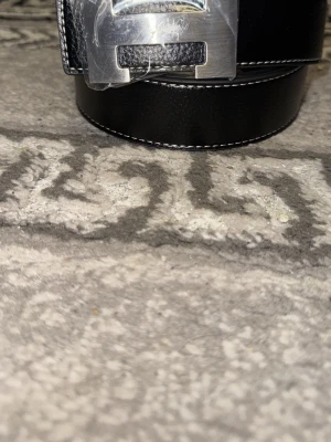 Svart skärp med silverfärgat spänne - Säljer ett stilrent svart skärp med kontrasterande vita sömmar och ett stort, silverfärgat metallspänne. Skärpet har en klassisk design och passar perfekt till jeans eller kostymbyxor. Materialet är slätt och ger en lyxig känsla.