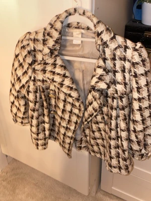 Rutig kavajjacka från Charlotte Russe - Säljer en snygg rutig trekvartsärmad kavajjacka från Charlotte Russe i beige och brunt. Jackan har stora guldknappar, bred krage och croppad passform. Materialmix med polyester, ull och bomull ger en lyxig känsla. Perfekt för dig som vill sticka ut med ett klassiskt men trendigt plagg.