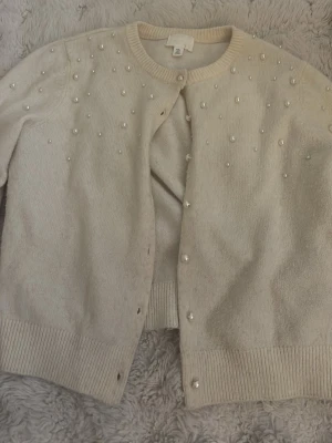 Beige kofta med pärlor från H&M - Supersöt beige kofta från H&M i storlek S. Koftan är dekorerad med pärlor på framsidan och har ribbade muddar vid ärmslut och nederkant. Stängs med knappar framtill och är perfekt för dig som gillar en mjuk och elegant look.