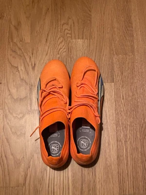 Puma Ultra orange fotbollsskor 44.5 - Säljer ett par grymt snygga Puma Ultra fotbollsskor i en riktigt skarp orange färg med blå och vita detaljer på sidan. Skorna har snörning och är tillverkade i syntetmaterial med dobbar för bästa grepp på planen. Perfekta för dig som vill sticka ut på matchen!