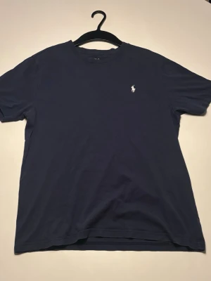 Marinblå t-shirt från Polo Ralph Lauren - Klassisk marinblå t-shirt från Polo Ralph Lauren med den ikoniska vita logon broderad på bröstet. T-shirten har rund hals och korta ärmar, tillverkad i mjuk bomull för en skön känsla. Perfekt till jeans eller shorts.
