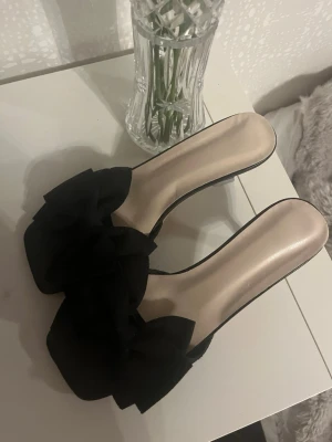 Svarta klackskor med rosett, strl 38 - Snygga svarta klackskor med öppen tå och stor rosett framtill. Skorna har låg klack och en stilren design. Perfekta för dig som vill ha något extra till outfiten🌸