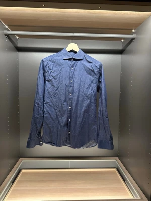 Blå slim fit skjorta från Riley - Snygg blå skjorta från Riley i slim fit-modell. Skjortan har diskreta vita prickar, klassisk krage och vita knappar framtill. Tillverkad i bomull och har långa ärmar med knapp vid manschetten. Perfekt för dig som gillar stilrena detaljer.