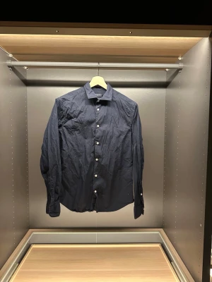 Mörkblå skjorta från Riley - Snygg mörkblå skjorta från Riley i slim fit-modell. Skjortan har klassisk krage, vita knappar framtill och långa ärmar. Tillverkad i ett lätt och skönt material som passar perfekt till många olika stilar.