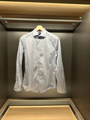 Ljusblå skjorta från Riley Slim Fit - Säljer en stilren ljusblå skjorta från Riley i slim fit-modell. Skjortan har klassisk krage, långa ärmar och knappar framtill. Tillverkad i mjuk bomull som känns skön mot huden. Perfekt för dig som gillar en clean och enkel look.