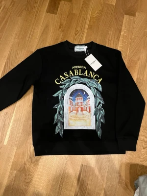 Svart sweatshirt från Casablanca - Säljer en svart sweatshirt från Casablanca med färgglad print framtill som föreställer ett fönster med arkitektur och blad. Tröjan har rund hals och långa ärmar. Perfekt för dig som vill ha något unikt och stilrent i garderoben.
