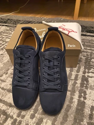 Mörkblå sneakers från Christian Louboutin - Snygga mörkblå sneakers från Christian Louboutin i mocka med klassisk röd sula. Skorna har rund tå, snörning och diskret broderad logga på hälen. Insidan är beige och materialet känns lyxigt och mjukt. Perfekta för dig som vill ha något exklusivt och stilrent.