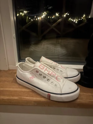 Vita BHPC sneakers med rosa tryck - Säljer ett par vita sneakers från BHPC med rosa logga och text på sidan. Skorna har metallfärgade öljetter, klassisk vit gummisula med svart rand och är tillverkade i bomullstyg. Perfekta för dig som gillar en clean och sportig look. Se skavanker på bilderna!