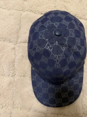Blå GG-mönstrad keps från Gucci - Säljer en snygg blå keps från Gucci med det klassiska GG-mönstret över hela kepsen. Justerbar rem bak i matchande blått och liten guldfärgad GG-detalj på sidan. Materialet är slitstark canvas med exklusiv känsla. Perfekt för dig som vill ha en statement-accessoar.
