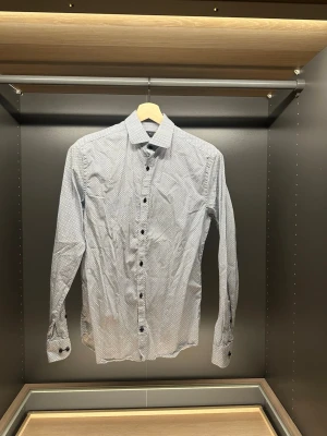 Slim fit skjorta från Riley, mönstrad - Säljer en vit långärmad skjorta från Riley med diskret geometriskt mönster i grått och blått. Skjortan har slim fit-passform, klassisk krage och svarta knappar. Perfekt för dig som gillar stilrena detaljer och snygg passform.