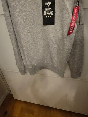 Grå sweatshirt från Alpha Industries - Snygg grå sweatshirt från Alpha Industries med klassisk passform. Tröjan har rund hals, ribbade muddar och en cool dragkedja på ärmen med röd 'Remove Before Flight'-tag. Perfekt för dig som gillar streetwear och vill ha en stilren look.