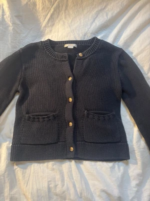 Mörkblå stickad kofta Gina Tricot - Mysig mörkblå stickad kofta från Gina Tricot med guldfärgade knappar och två fickor framtill. Klassisk rund halsringning och ribbade muddar vid ärmslut och nederkant. Perfekt för lager på lager under kyliga dagar.