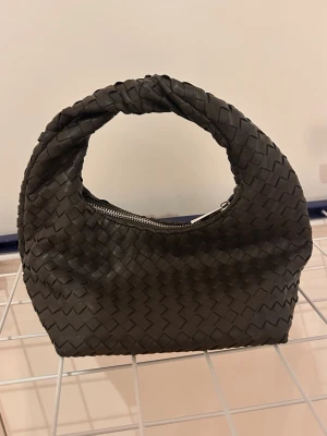 Flätad brun handväska i skinn - Säljer en snygg brun handväska i flätat skinn med rundat handtag och dragkedja upptill. Väskan har en modern, chunky look och är perfekt för dig som gillar trendiga accessoarer. Enkel att matcha till olika outfits och rymmer det viktigaste.