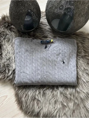 Grå kabelstickad tröja Polo Ralph Lauren - Storlek: M - Säljer en grå kabelstickad tröja från Polo Ralph Lauren. Tröjan har rund halsringning, lång ärm och den klassiska broderade loggan på bröstet. Perfekt för lager-på-lager och snygg till jeans. Storlek: M