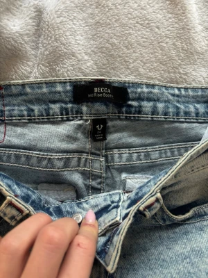 True Religion Becca Mid Rise Bootcut w24 Jeans - Jag säljer ett par ljusblå True Religion Becca jeans med bootcut  midwaist i w24. Jeansen har hål vid knäna, klassiska bakfickor med broderad logga och detaljer i rött. Tillverkade i jeansmaterial med en relaxed passform. Har använt jeansen inte så mycket för de blev för små för mig🫰🏻❤️