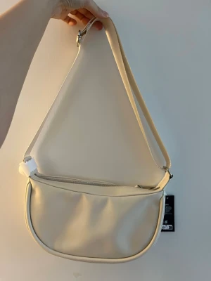 Beige axelväska i syntet - Snygg beige axelväska med minimalistisk design. Väskan har en justerbar axelrem och stängs med dragkedja upptill. Tillverkad i skinn material och har en stilren, rundad form som passar till många olika looks.