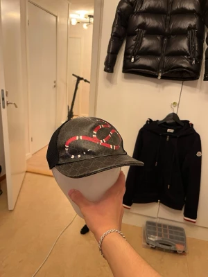 Gucci keps - Säljer en ÄKTA svart Gucci keps (kingsnake). Säljer den då ja ej använder. Kepsen kommer med box dustbag kvitto och alla tillbehör/tags. Kepsen är som ny ända nackdelen är att vid spännbandet ibak har en liten bit av lädret som är på räderna gått sönder, men inget större problem då de knappast syns. Kepsen säljs inte heller på Guccis hemsida längre.
