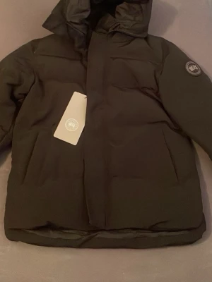 Canada goose Macmillan - Säljer en svart pufferjacka från Canada Goose med huva och diskret logga på ärmen. Jackan har en stilren design med raka linjer och stora fickor framtill. Perfekt för kalla dagar och riktigt stilig. Jackan är vadderad och har en klassisk passform.